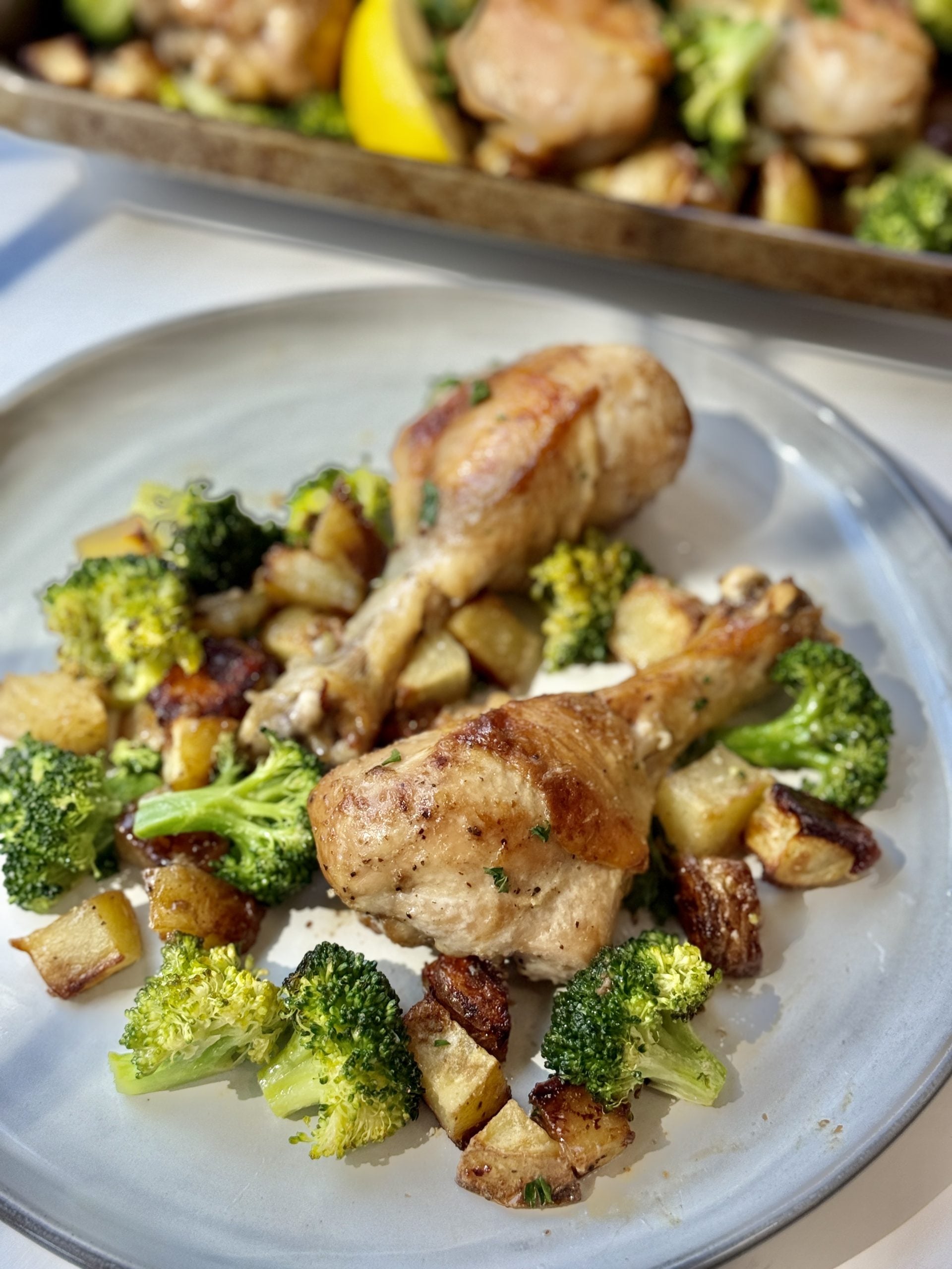 A plate with two roasted chicken drumsticks, surrounded by roasted broccoli and diced potatoes, with a baking tray of more chicken and veggies in the background.