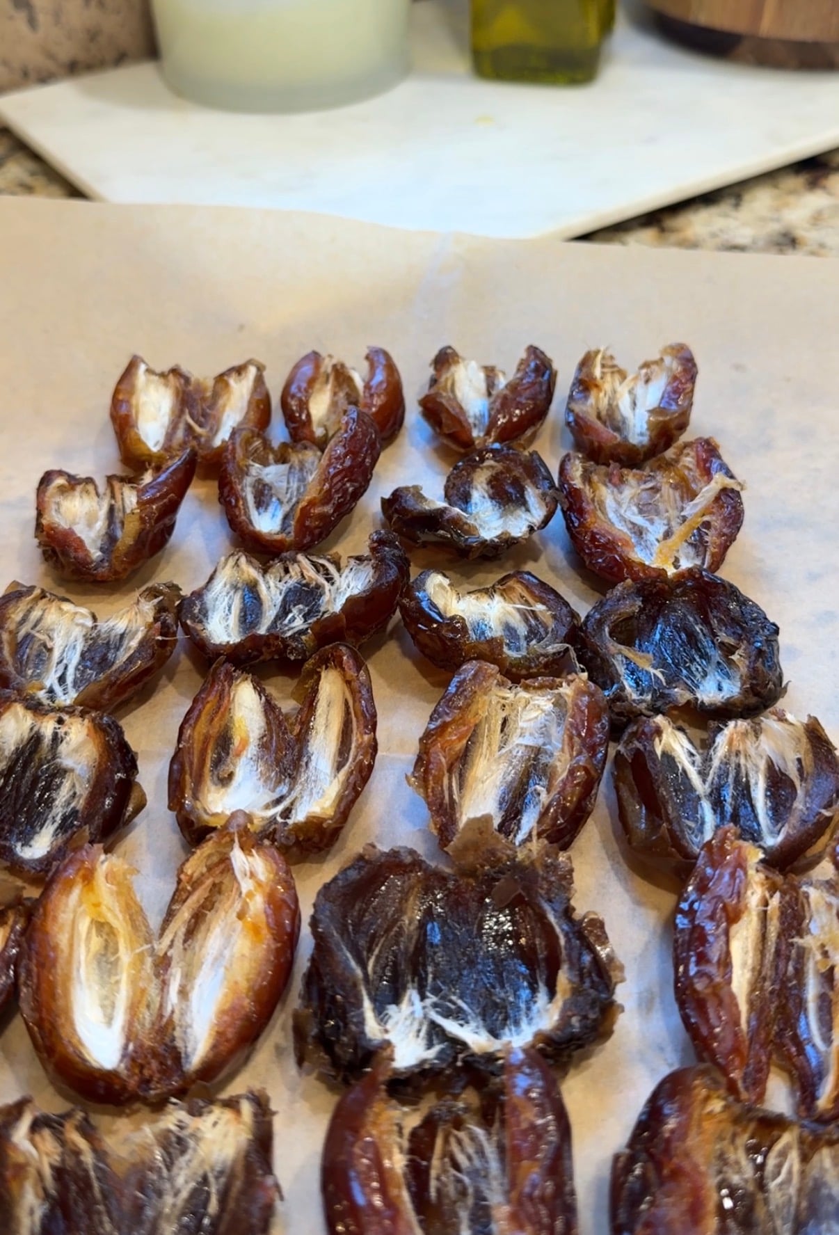 Halved dates with pits removed are arranged on a sheet of parchment paper, ready for preparation, on a kitchen countertop.