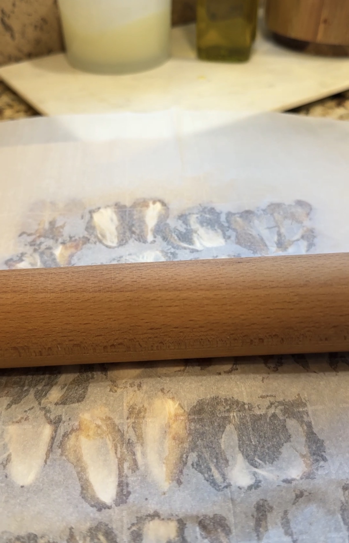 A wooden rolling pin on parchment paper, pressing down on garlic cloves, partially flattening them on a speckled countertop.