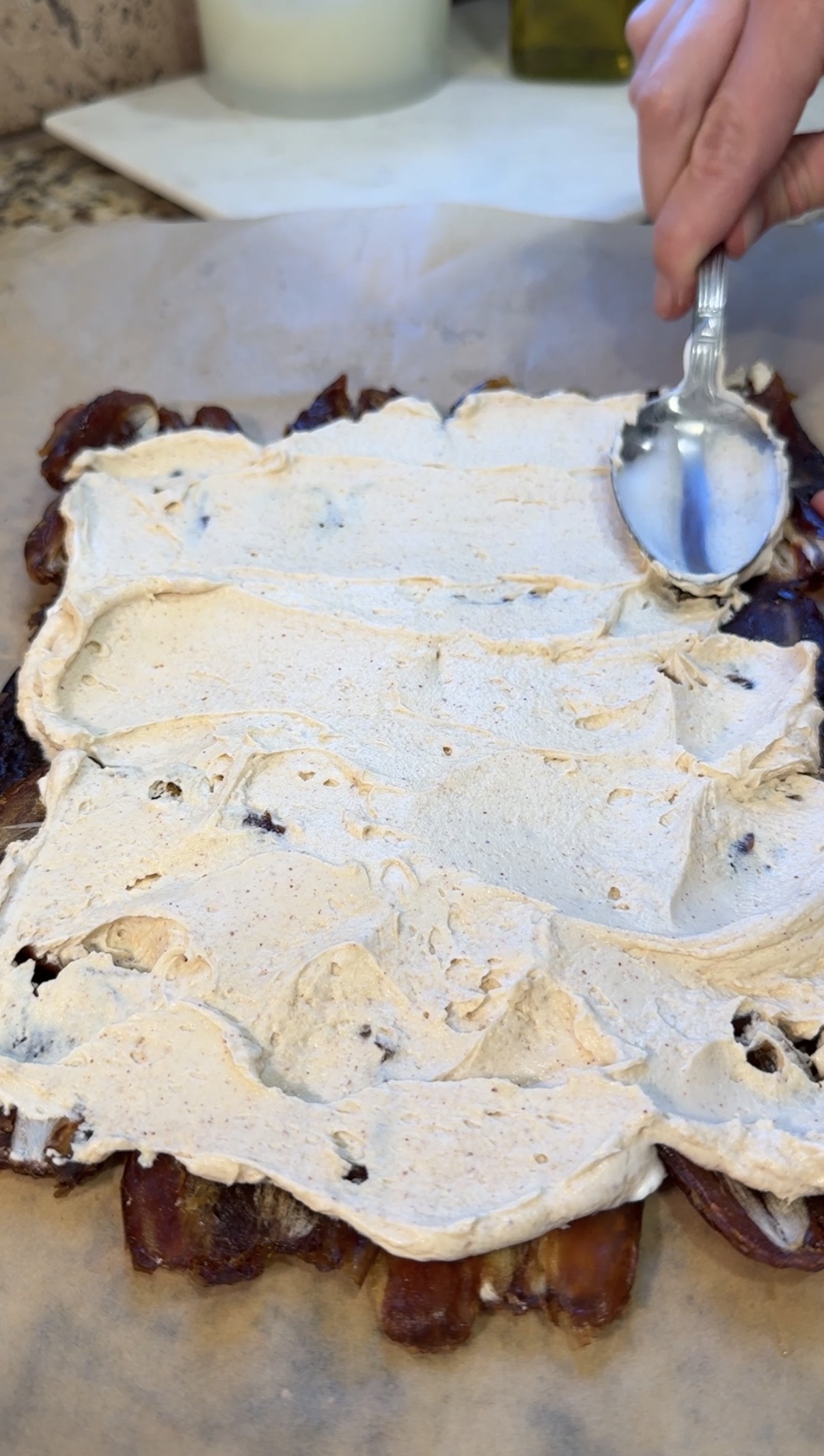 A hand uses a spoon to spread a creamy, light brown mixture over a square layer of bacon strips on parchment paper.