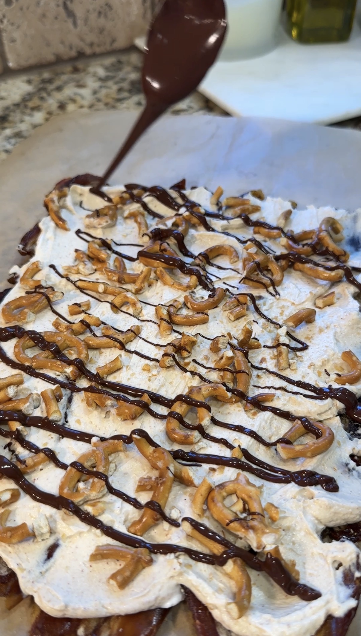 A dessert topped with whipped cream, crushed pretzels, and chocolate drizzle is being finished with more chocolate sauce from a spoon.