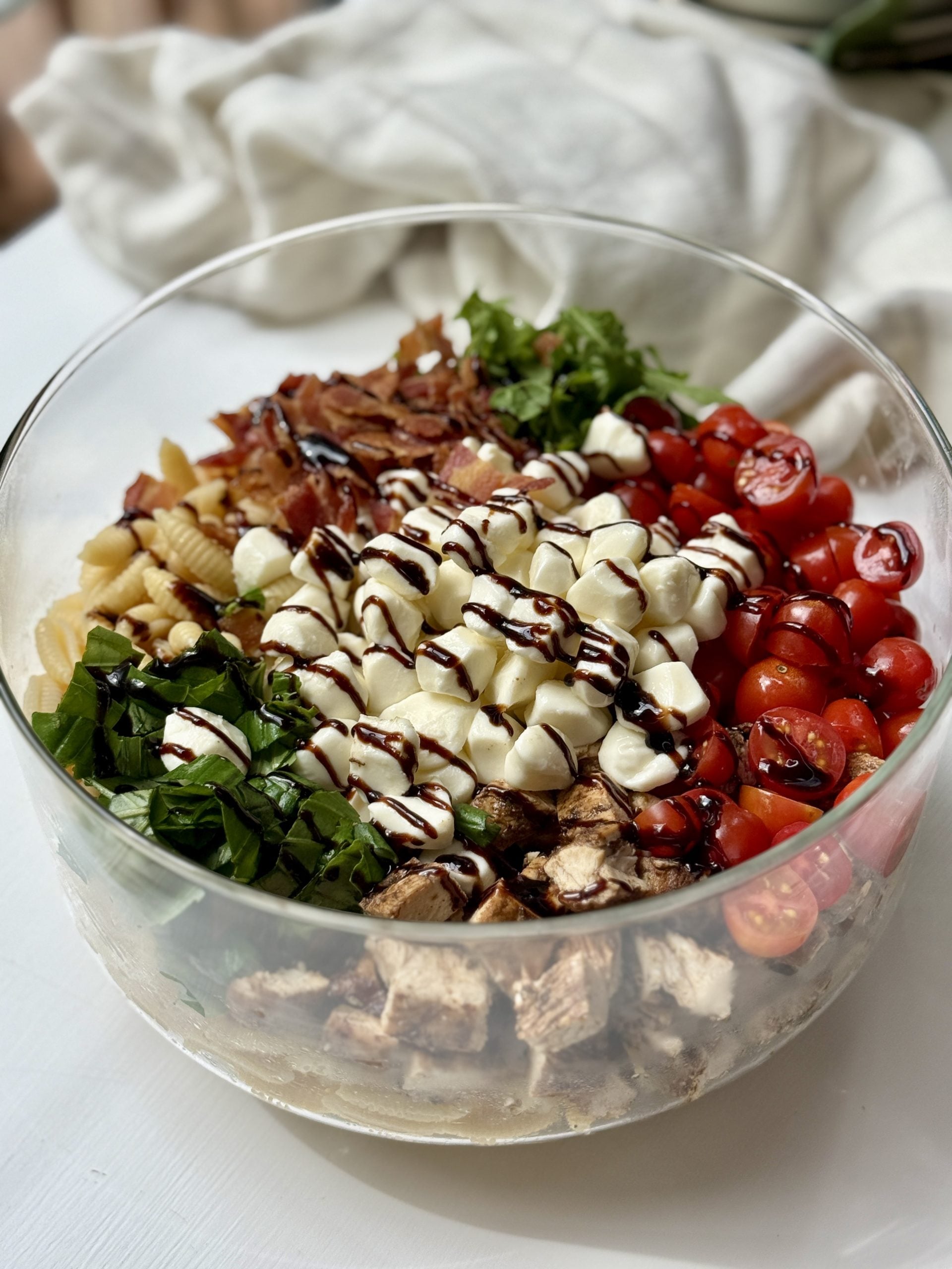 A glass bowl filled with colorful ingredients: cooked pasta, chopped greens, grilled chicken, cherry tomatoes, mozzarella balls, bacon, and a balsamic glaze drizzle, arranged in sections on a white surface.