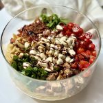 A glass bowl filled with colorful ingredients: cooked pasta, chopped greens, grilled chicken, cherry tomatoes, mozzarella balls, bacon, and a balsamic glaze drizzle, arranged in sections on a white surface.