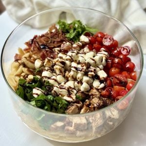 A glass bowl filled with colorful ingredients: cooked pasta, chopped greens, grilled chicken, cherry tomatoes, mozzarella balls, bacon, and a balsamic glaze drizzle, arranged in sections on a white surface.