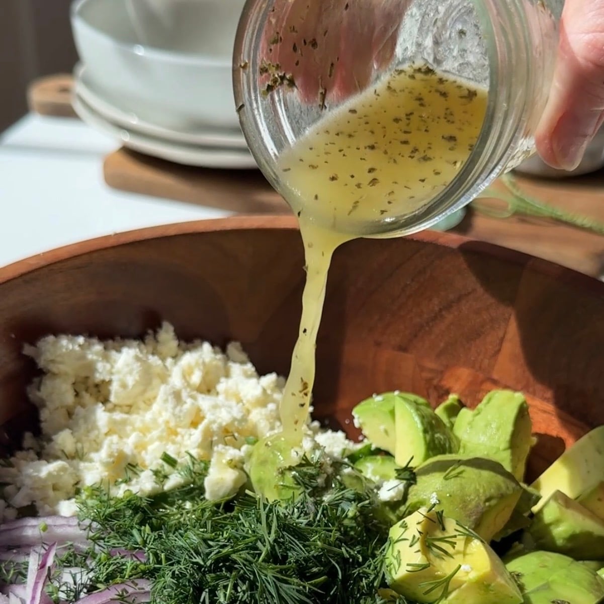 A close-up of a wooden bowl filled with chopped avocado, sliced red onion, crumbled cheese, fresh dill, and dressing being poured on top.