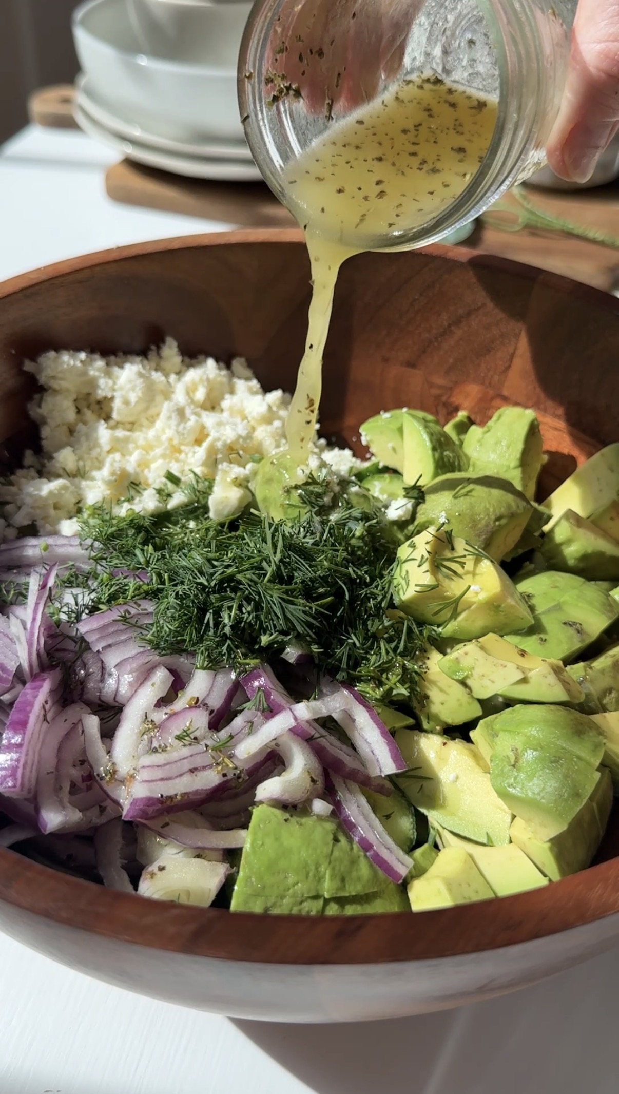 A close-up of a wooden bowl filled with chopped avocado, sliced red onion, crumbled cheese, fresh dill, and dressing being poured on top.