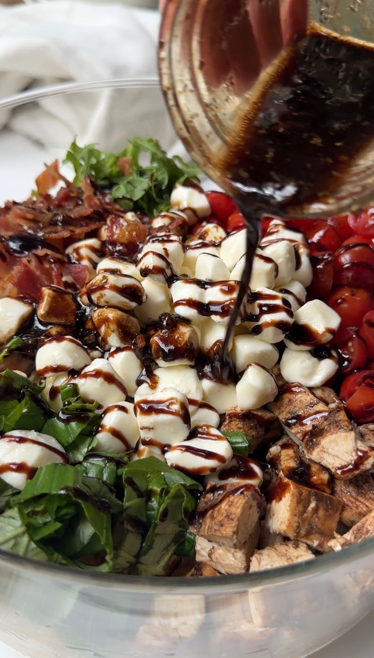 A close-up of a fresh salad with chopped greens, cherry tomatoes, mozzarella balls, bacon, and grilled chicken, as balsamic dressing is being poured over the top.
