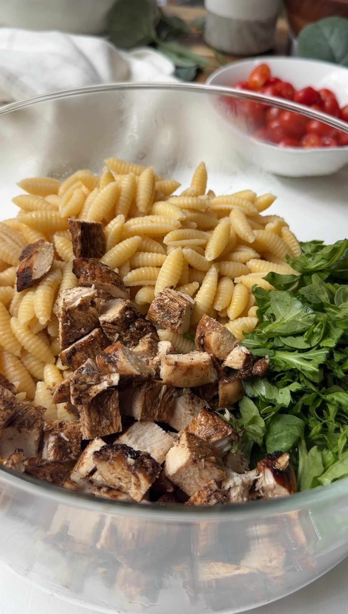 A glass bowl filled with cooked pasta, chopped grilled chicken, and fresh green spinach, with a bowl of cherry tomatoes in the background.