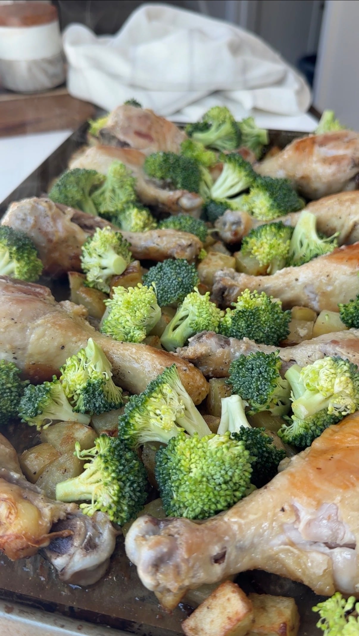 A baking tray filled with roasted chicken drumsticks, broccoli florets, and diced potatoes, all cooked and lightly browned.