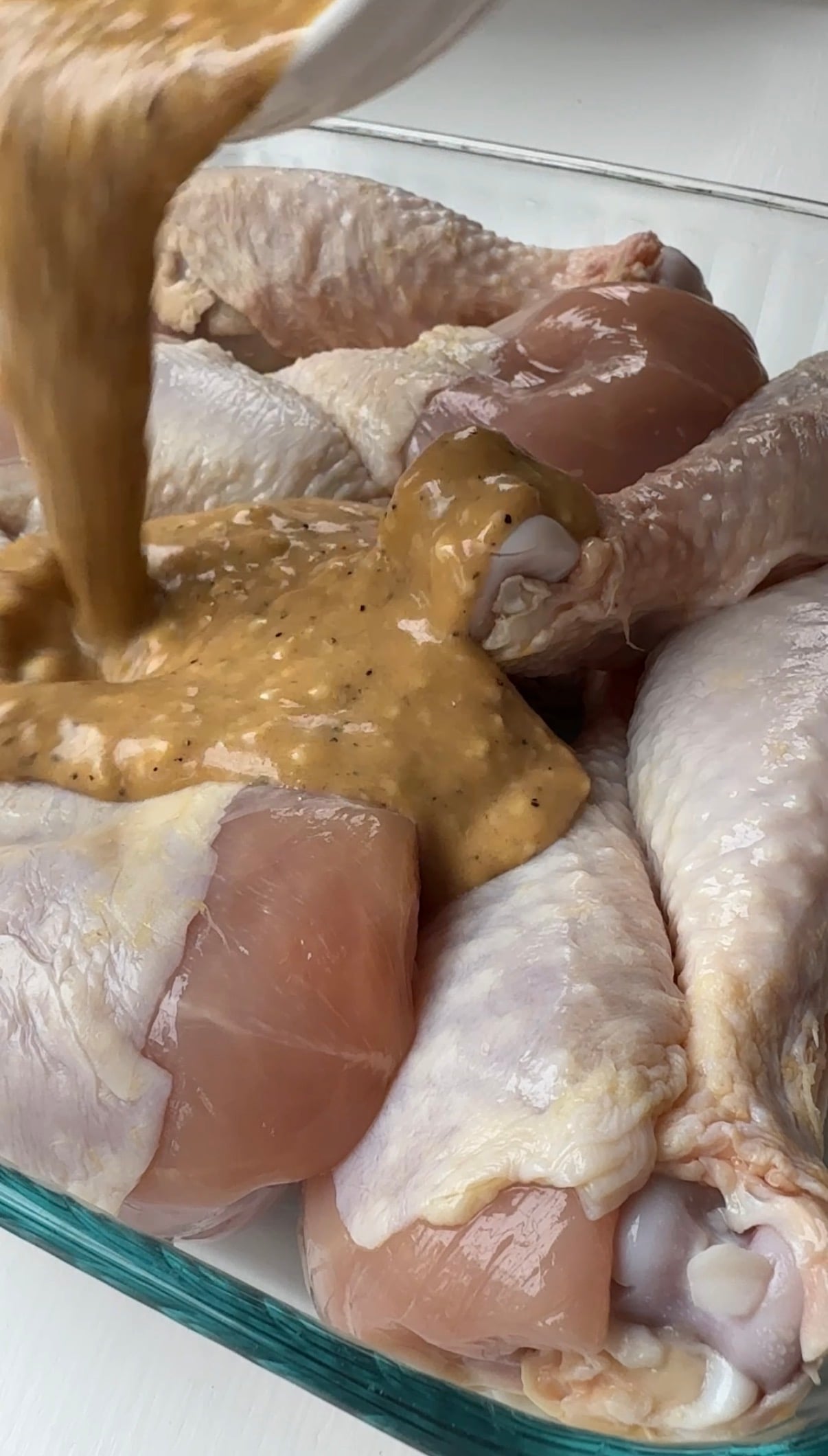 Close-up of raw chicken drumsticks and thighs in a glass dish, with a thick, seasoned marinade being poured over them.