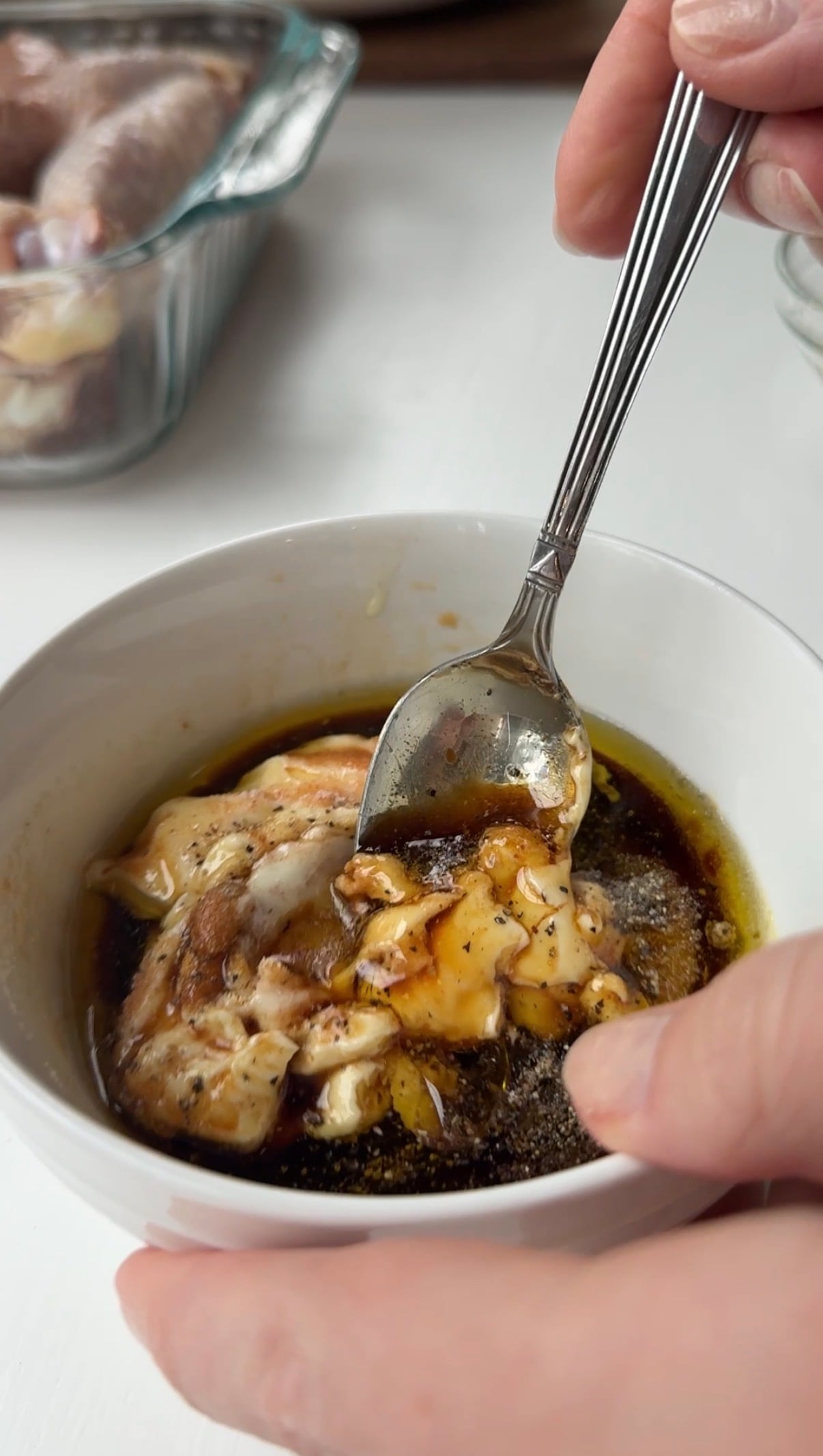 A hand holds a spoon mixing honey, mustard, and spices in a white bowl with oil and soy sauce, preparing a marinade. In the background, raw chicken pieces are visible in a glass dish.