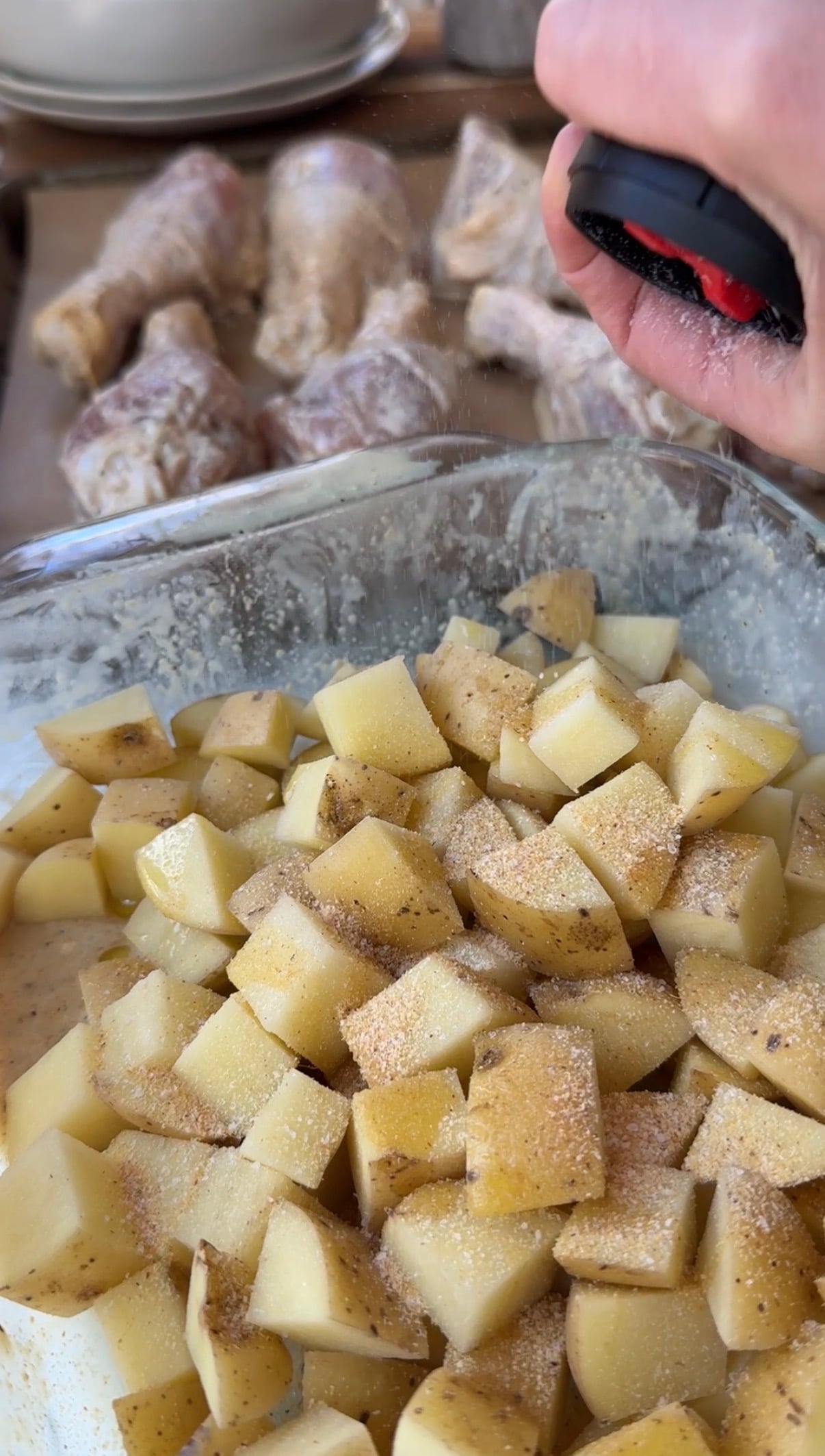 A hand sprinkles seasoning over a glass dish filled with cubed potatoes. In the background, uncooked, seasoned chicken drumsticks are visible on a tray.
