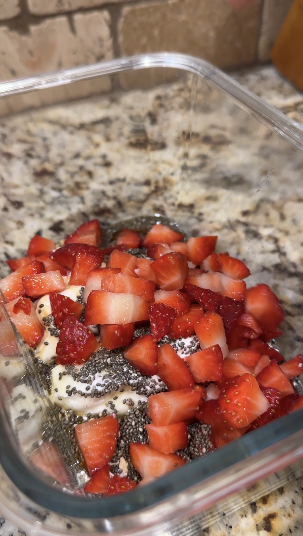 A clear glass container on a granite countertop is filled with chopped fresh strawberries, chia seeds, and what appears to be yogurt or oatmeal.