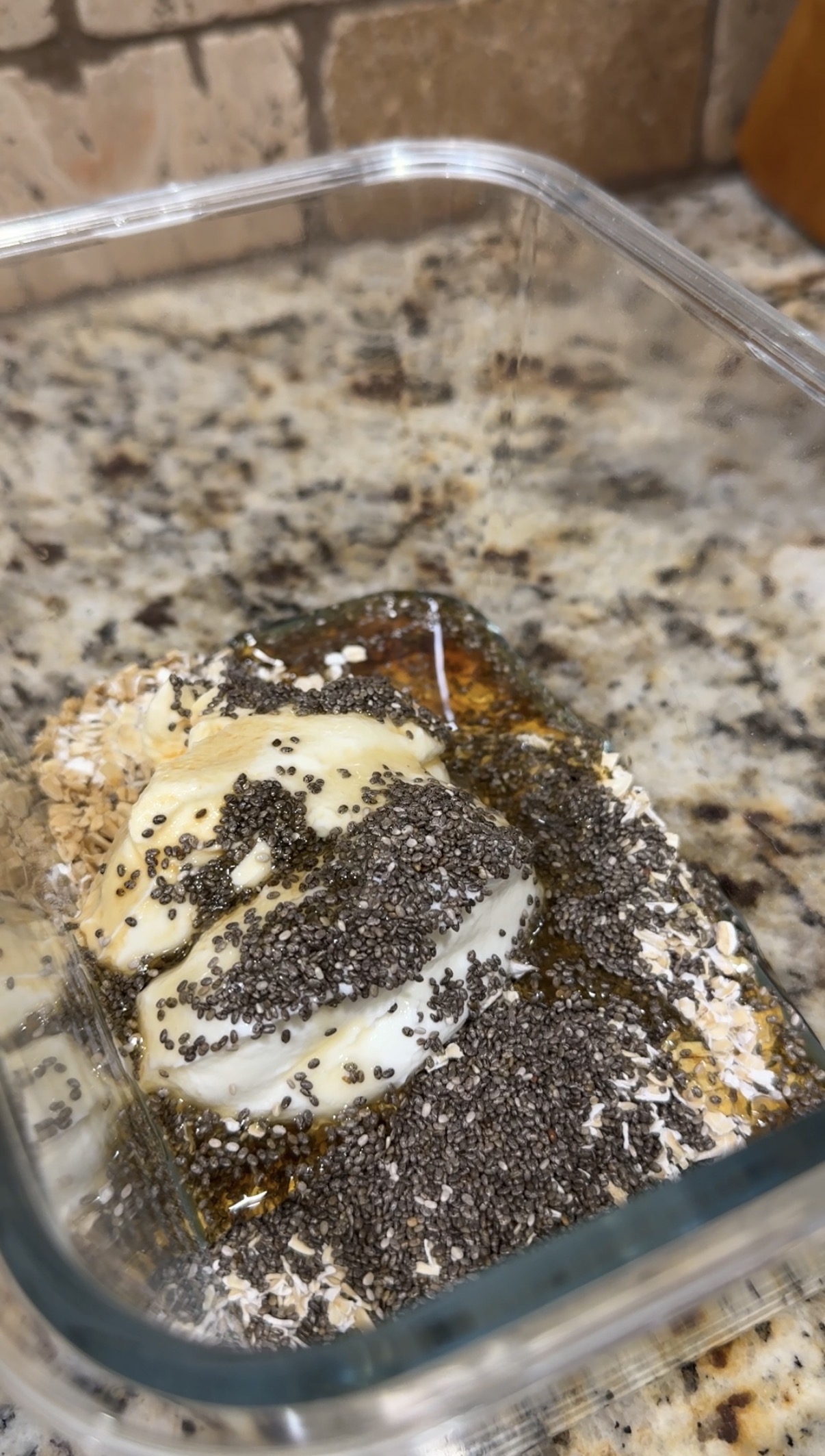 A glass container on a granite countertop holds oats, Greek yogurt, chia seeds, honey, and peanut butter, ready to be mixed for an overnight oats recipe.