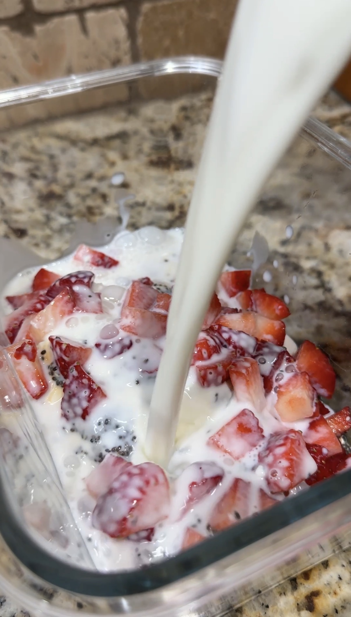 Milk being poured over chopped strawberries and chia seeds in a glass container, with a granite countertop in the background.