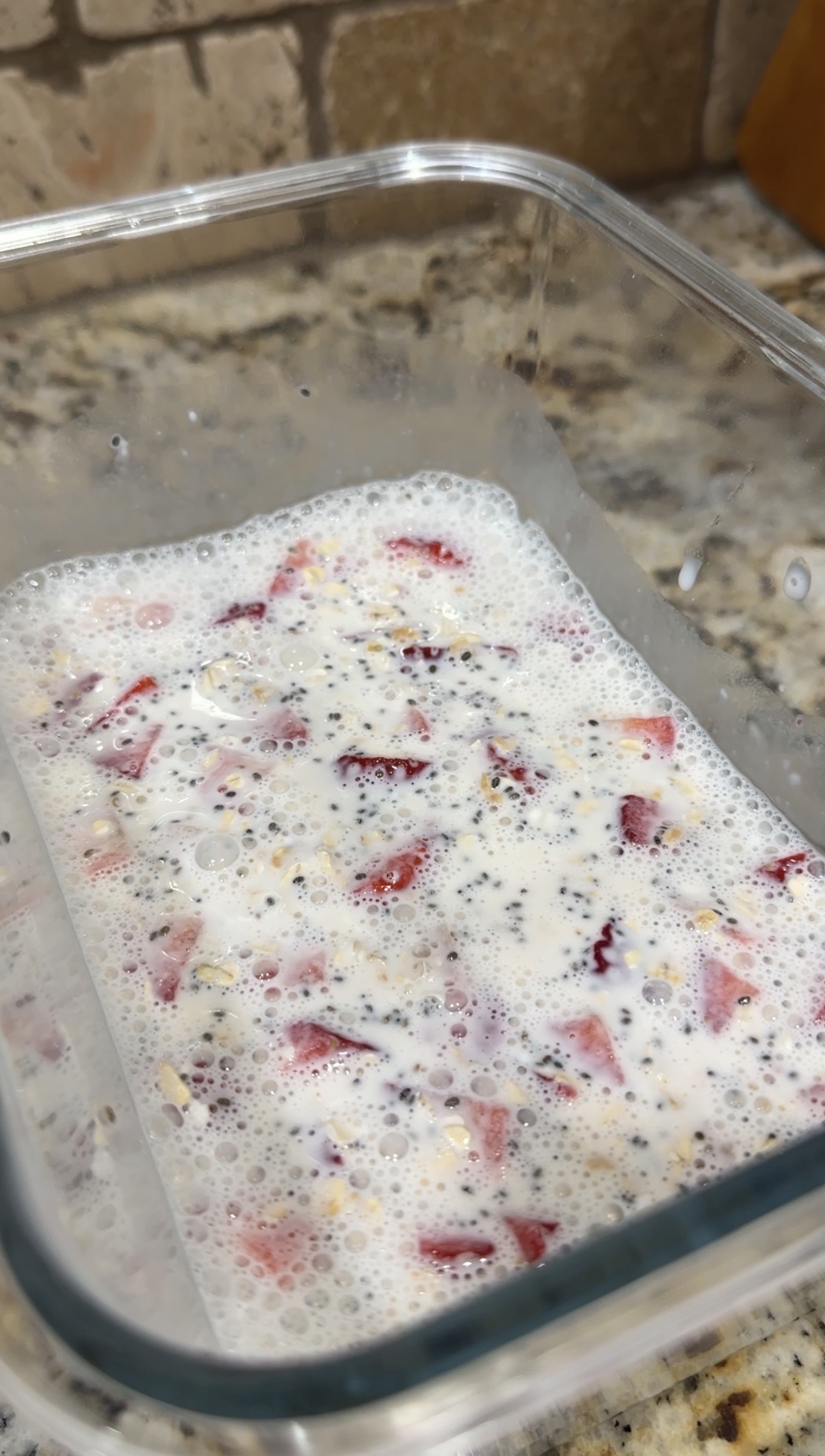A close-up of a glass container filled with a mixture of milk, chopped strawberries, chia seeds, and oats, sitting on a speckled kitchen countertop.