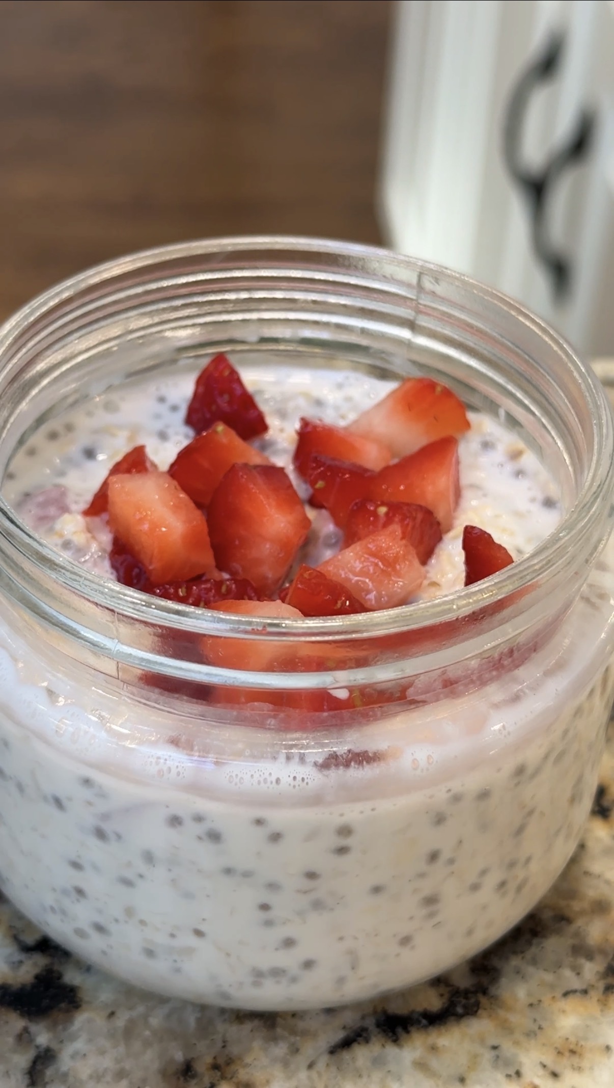 A glass jar filled with chia seed pudding, topped with chopped fresh strawberries, sits on a speckled countertop. The pudding appears creamy with visible chia seeds throughout.