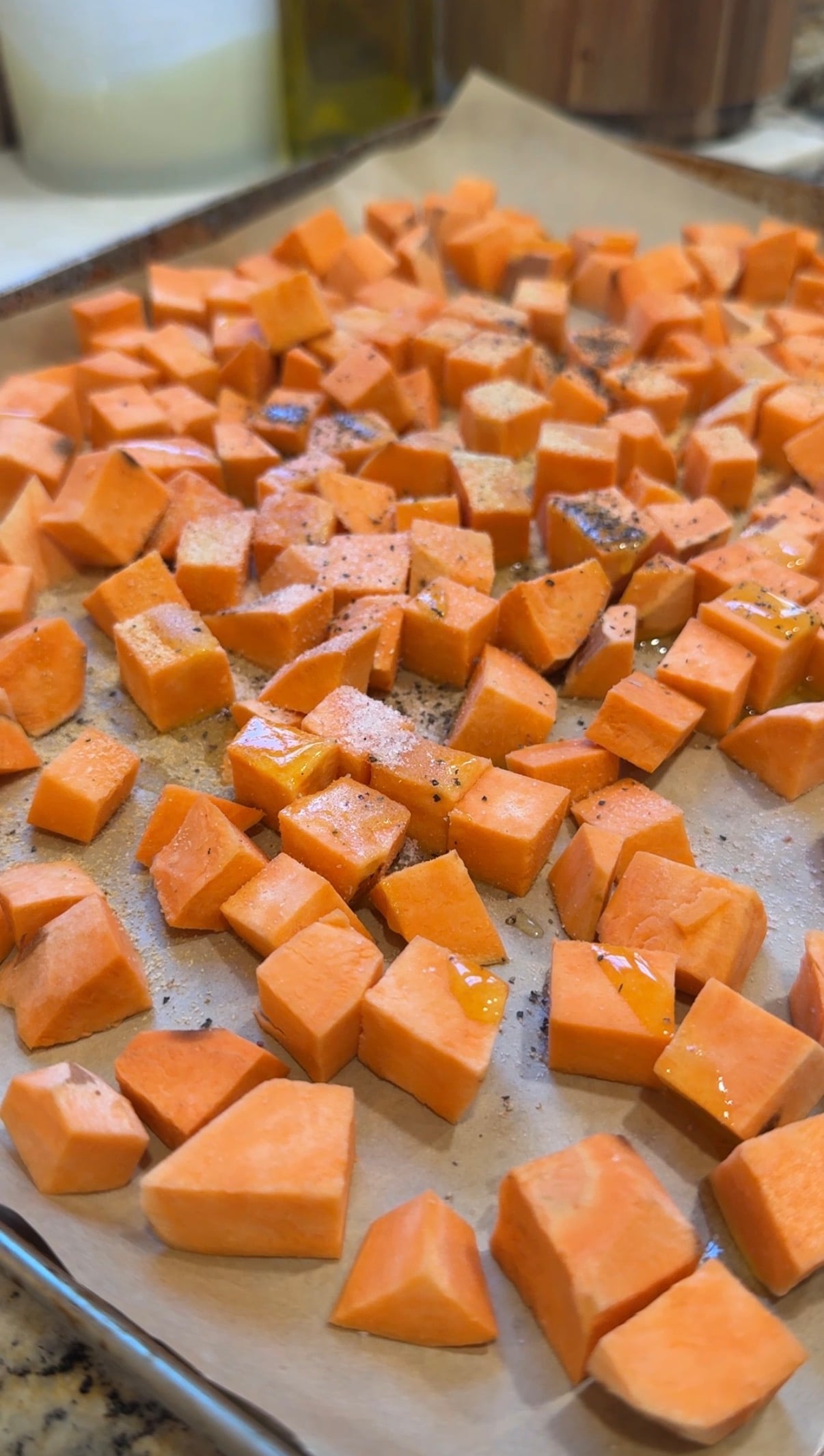 Diced sweet potatoes spread on a parchment-lined baking sheet, drizzled with oil and sprinkled with ground black pepper, ready for roasting.