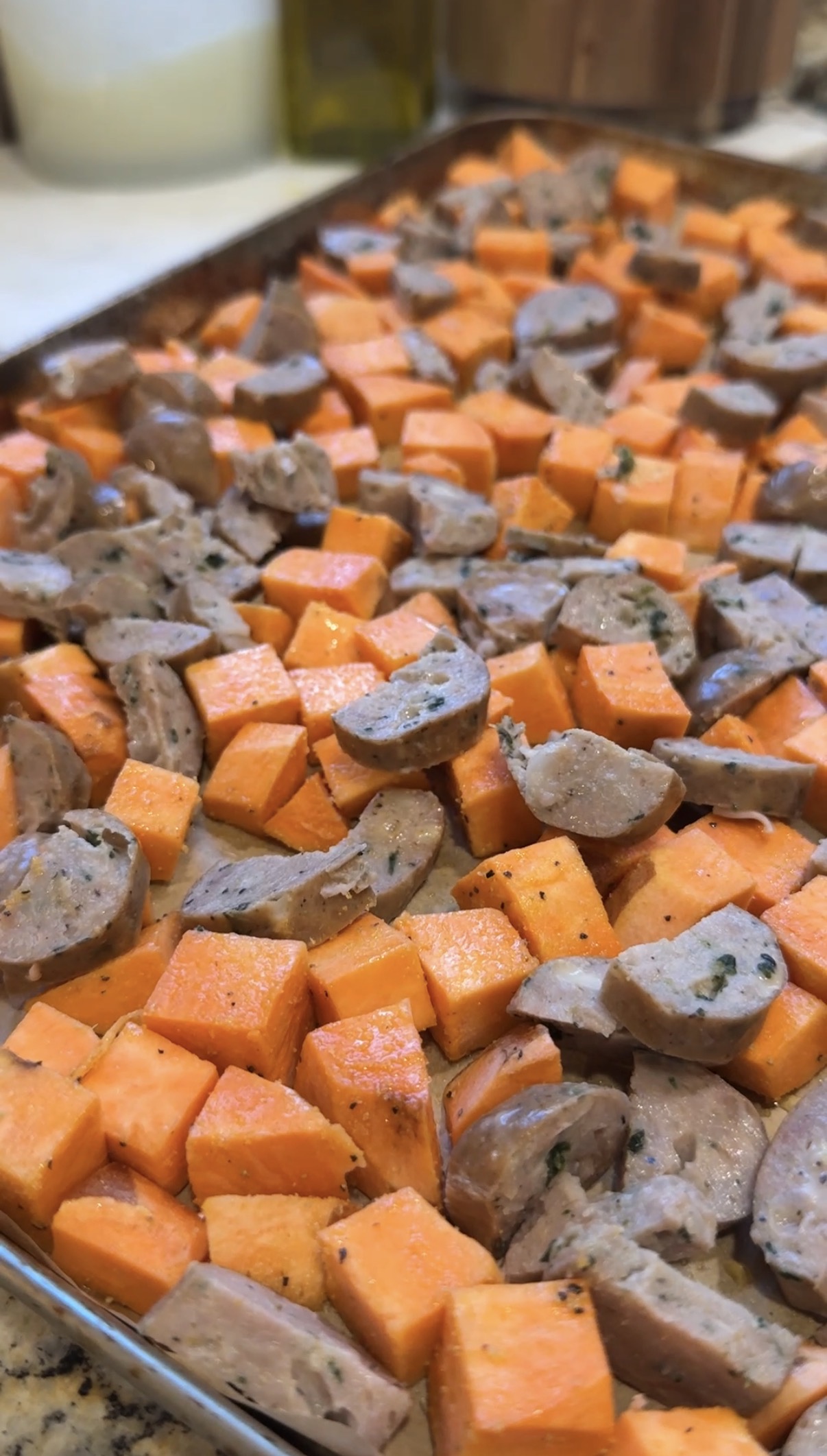 Diced sweet potatoes spread on a parchment-lined baking sheet, drizzled with oil and sprinkled with ground black pepper, ready for roasting.