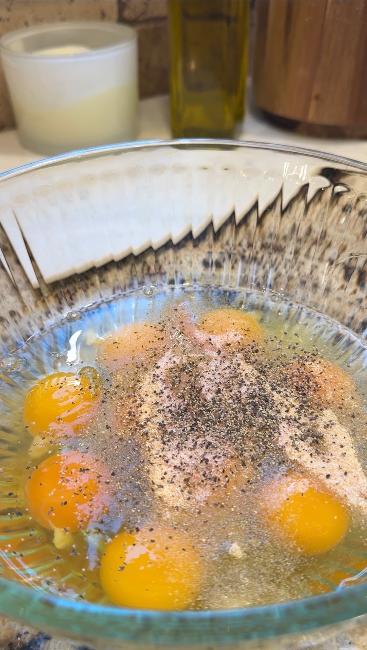 A glass bowl containing cracked eggs, seasoning, and black pepper on top, sitting on a kitchen counter with a candle, olive oil bottle, and wooden container in the background.