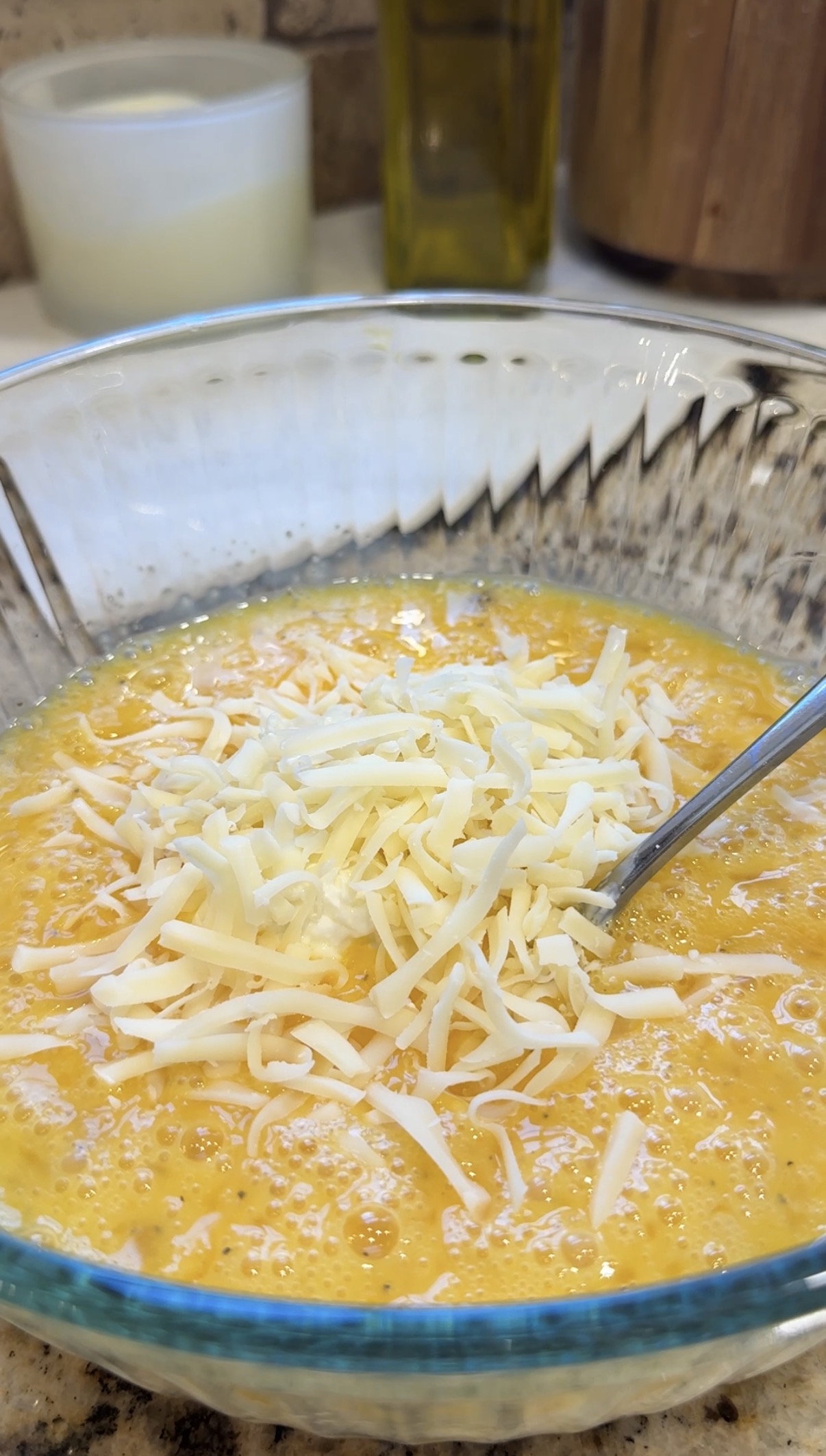 A glass bowl filled with beaten eggs topped with shredded cheese, with a metal fork resting inside.