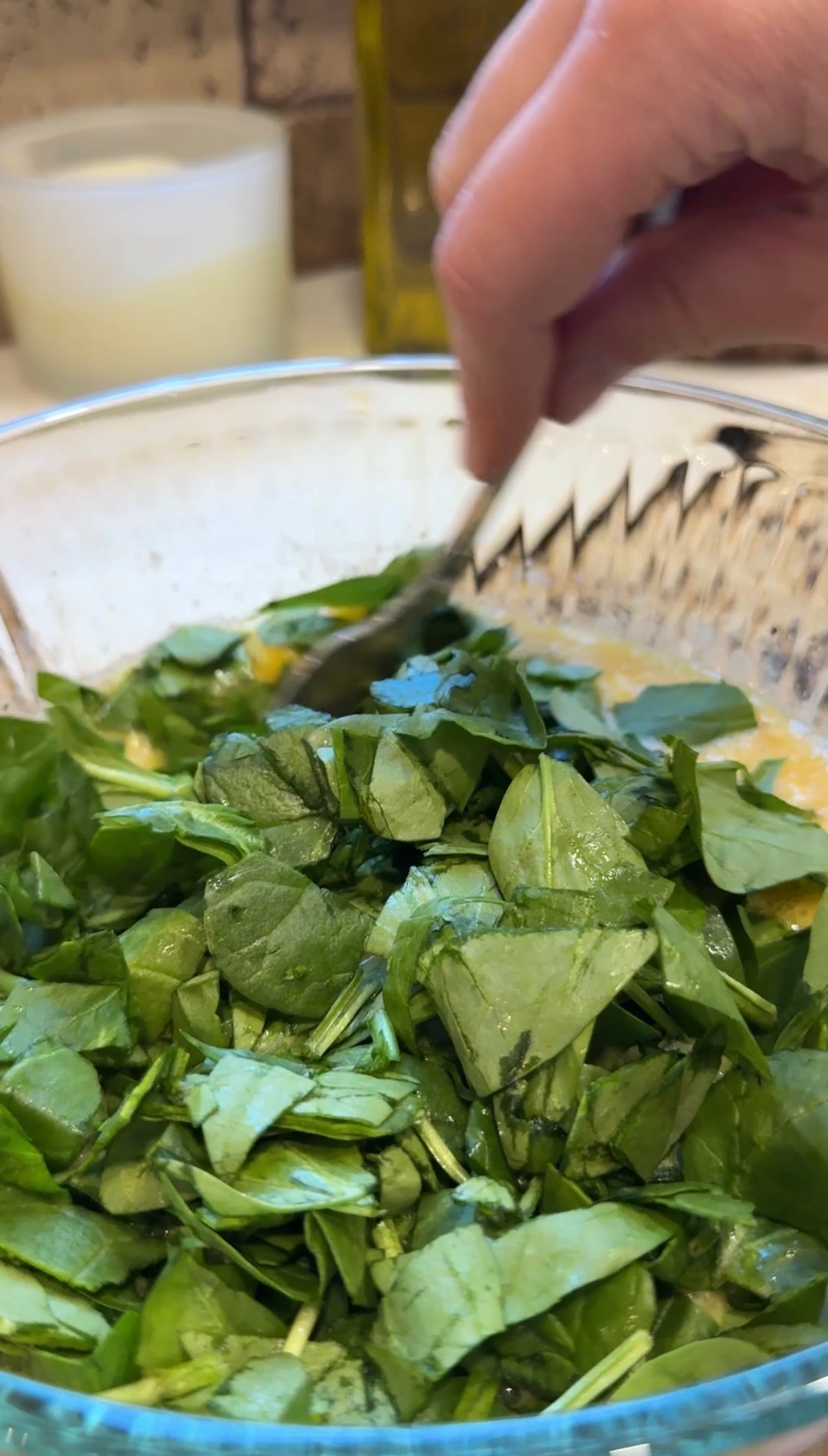 A hand stirs chopped fresh spinach leaves in a glass bowl containing a light-colored liquid mixture, possibly preparing a salad or a spinach-based dish.