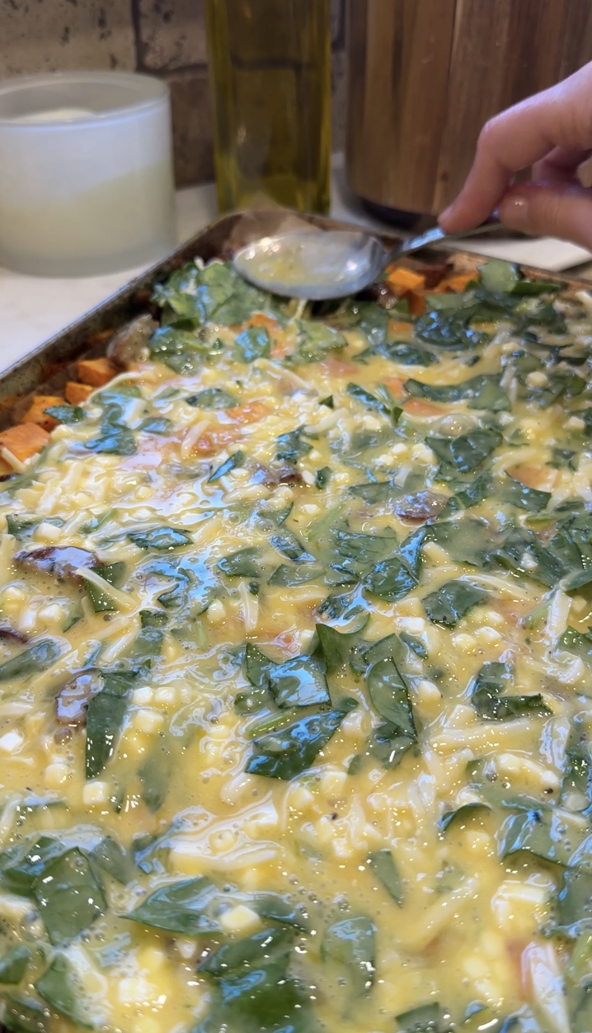 A close-up of an unbaked egg casserole in a baking dish, filled with chopped greens, cheese, diced sweet potatoes, and other vegetables, with a spoon held over the mixture.