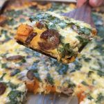 A close-up of a spatula lifting a slice of baked casserole topped with melted cheese, sliced sausage, spinach, and orange vegetables, with the remaining casserole visible in the background.