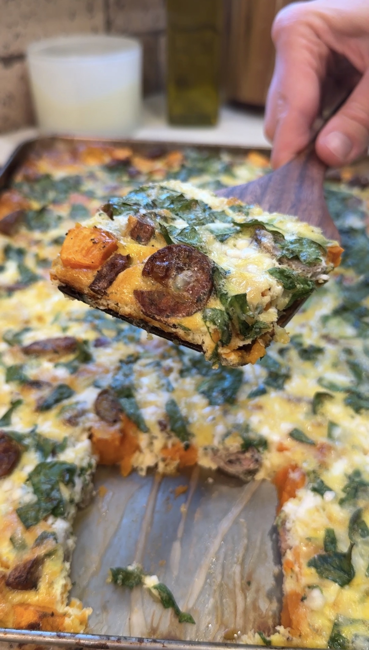 A close-up of a spatula lifting a slice of baked casserole topped with melted cheese, sliced sausage, spinach, and orange vegetables, with the remaining casserole visible in the background.