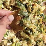 A hand holds a chip scooping up a chunky salad mix with lettuce, avocado, and creamy dressing on a wooden surface.