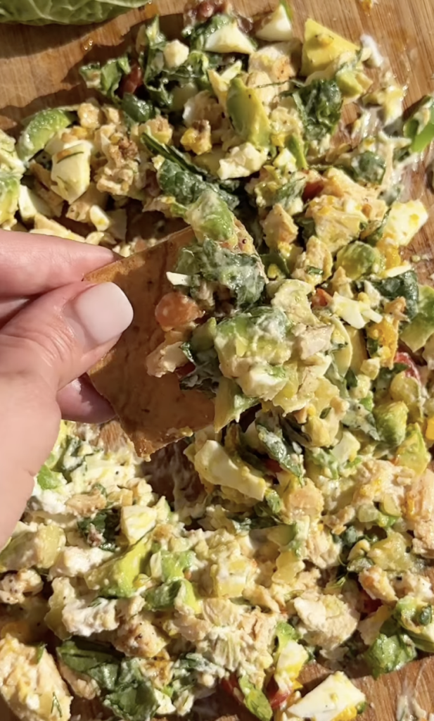 A hand holds a chip scooping up a chunky salad mix with lettuce, avocado, and creamy dressing on a wooden surface.