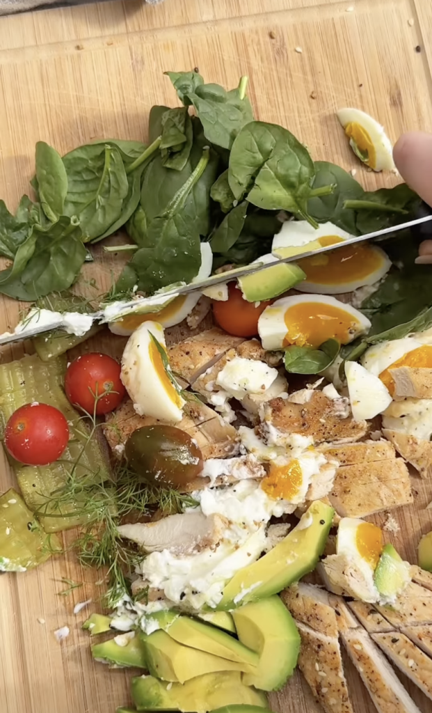 A cutting board with fresh spinach, cherry tomatoes, sliced avocado, pickles, soft-boiled eggs with runny yolks, grilled chicken, and a sprinkling of herbs. A hand holds a knife, slicing through the food.