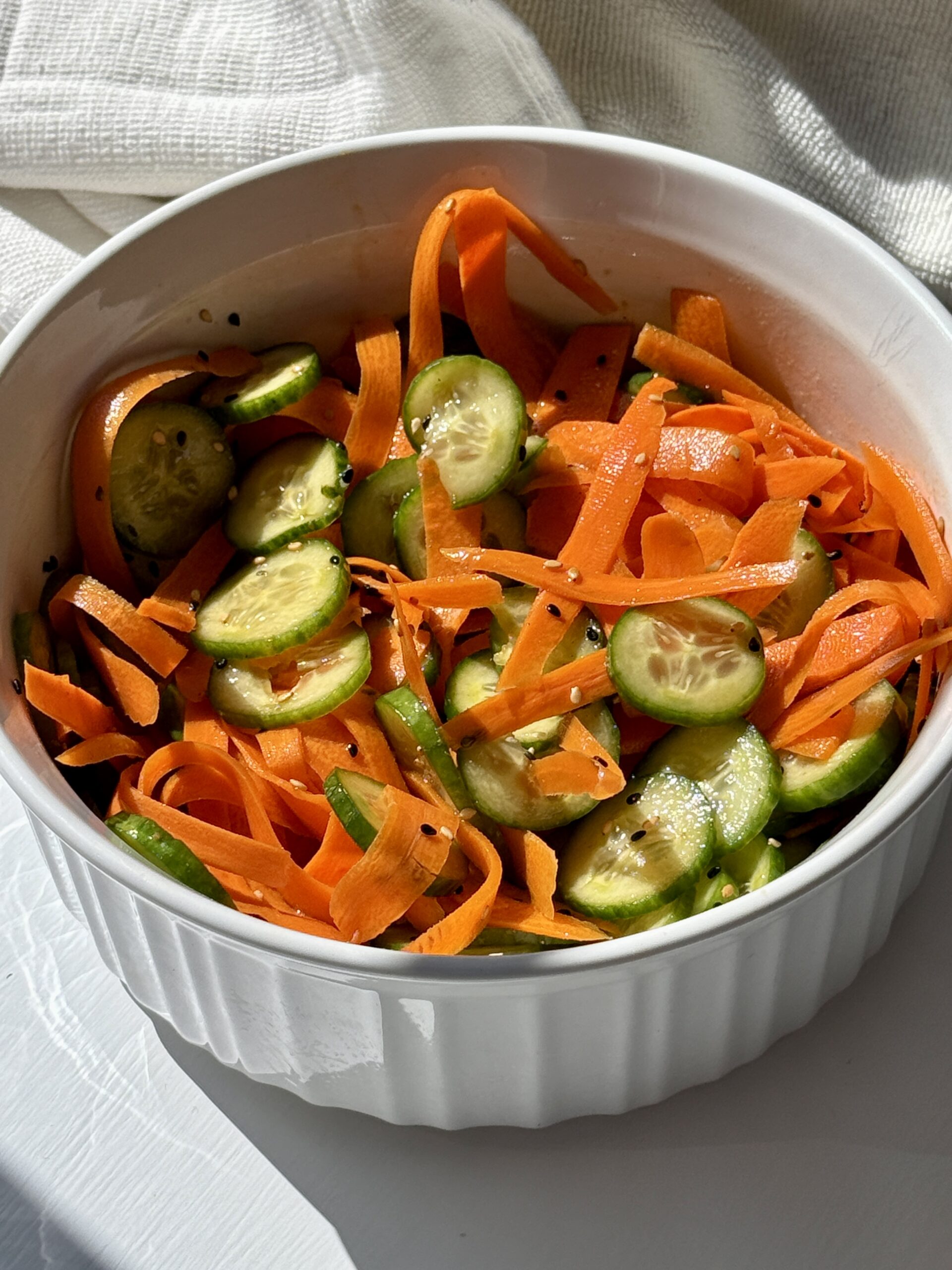 A white bowl filled with a fresh salad of sliced cucumbers and thinly shaved carrot ribbons, lightly seasoned, sits on a white surface in natural sunlight.