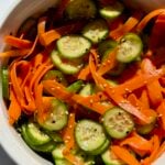 A white bowl filled with a fresh salad of sliced cucumbers and thinly shaved carrot ribbons, lightly seasoned, sits on a white surface in natural sunlight.