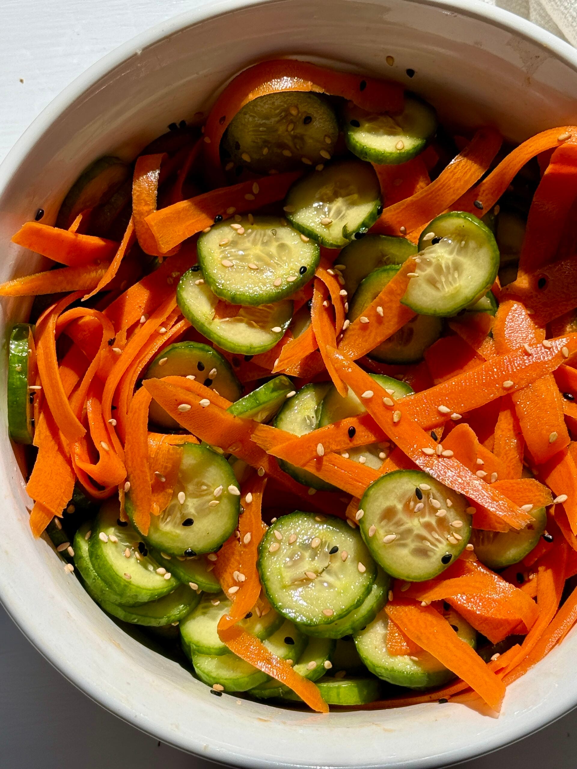 A white bowl filled with a fresh salad of sliced cucumbers and thinly shaved carrot ribbons, lightly seasoned, sits on a white surface in natural sunlight.