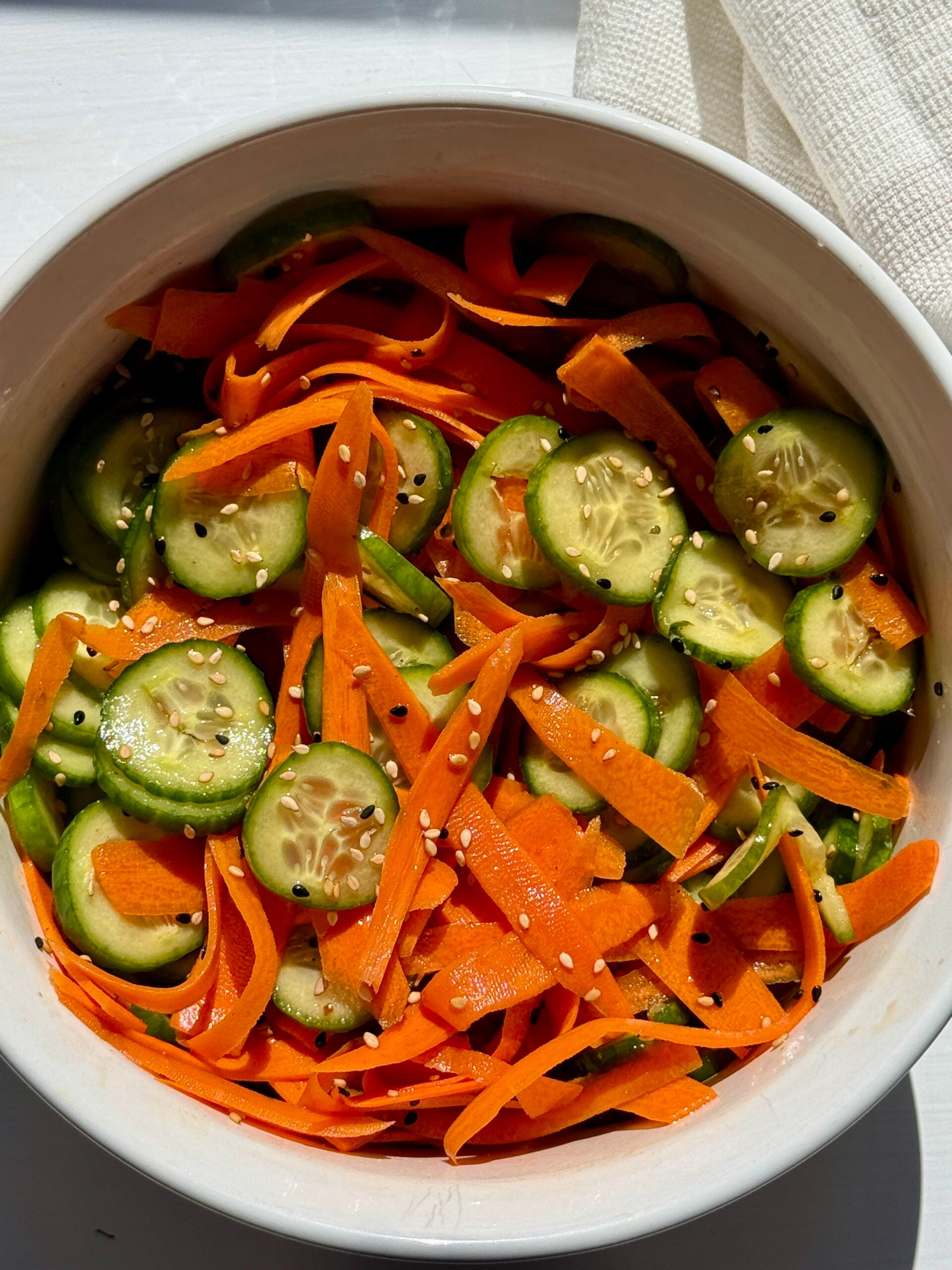 A white bowl filled with a fresh salad of sliced cucumbers and thinly shaved carrot ribbons, lightly seasoned, sits on a white surface in natural sunlight.