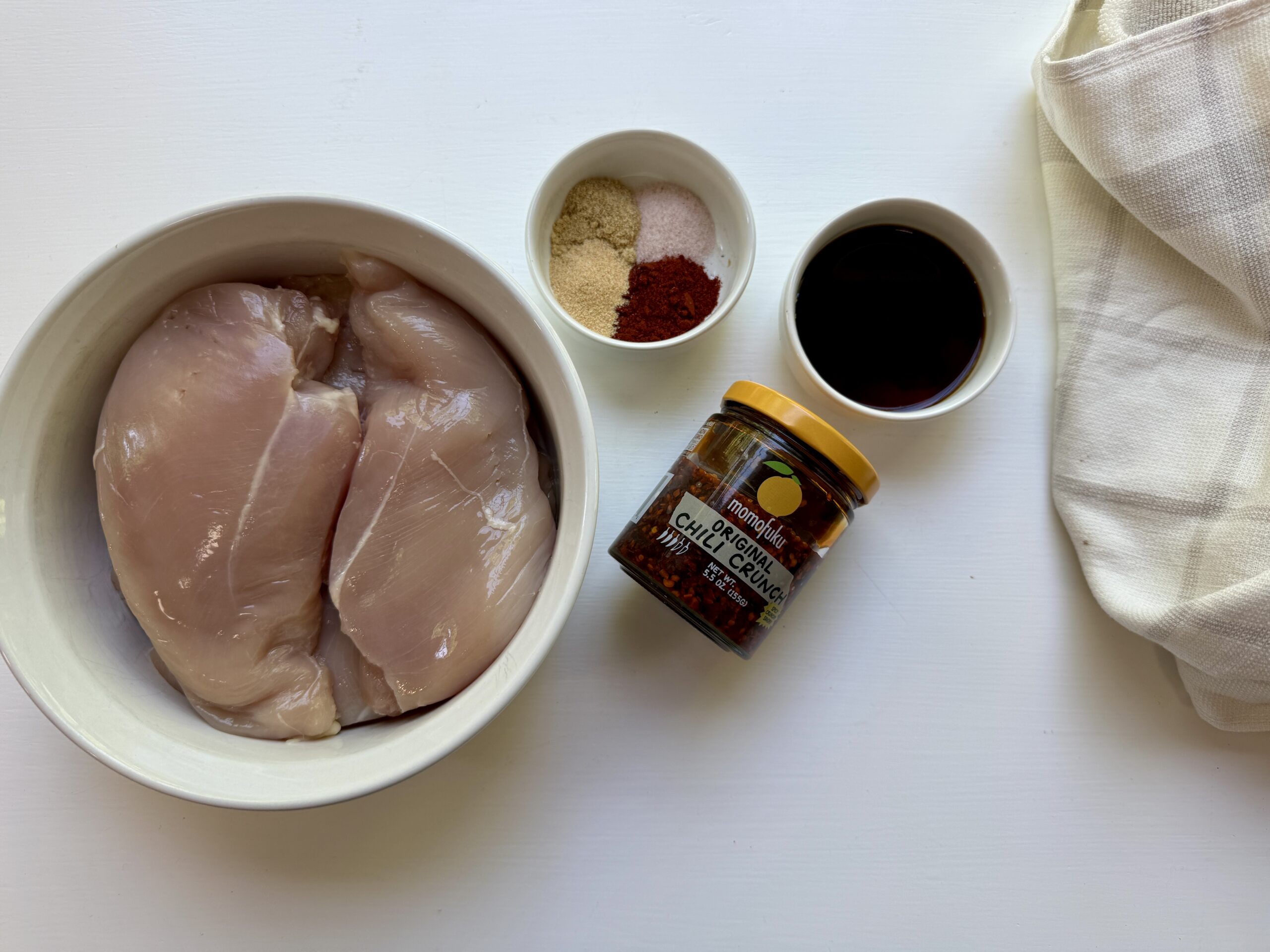 Two raw chicken breasts in a white bowl, with small bowls of spices, a jar of chipotle chile, and a small bowl of dark liquid arranged on a white surface beside a folded white cloth.