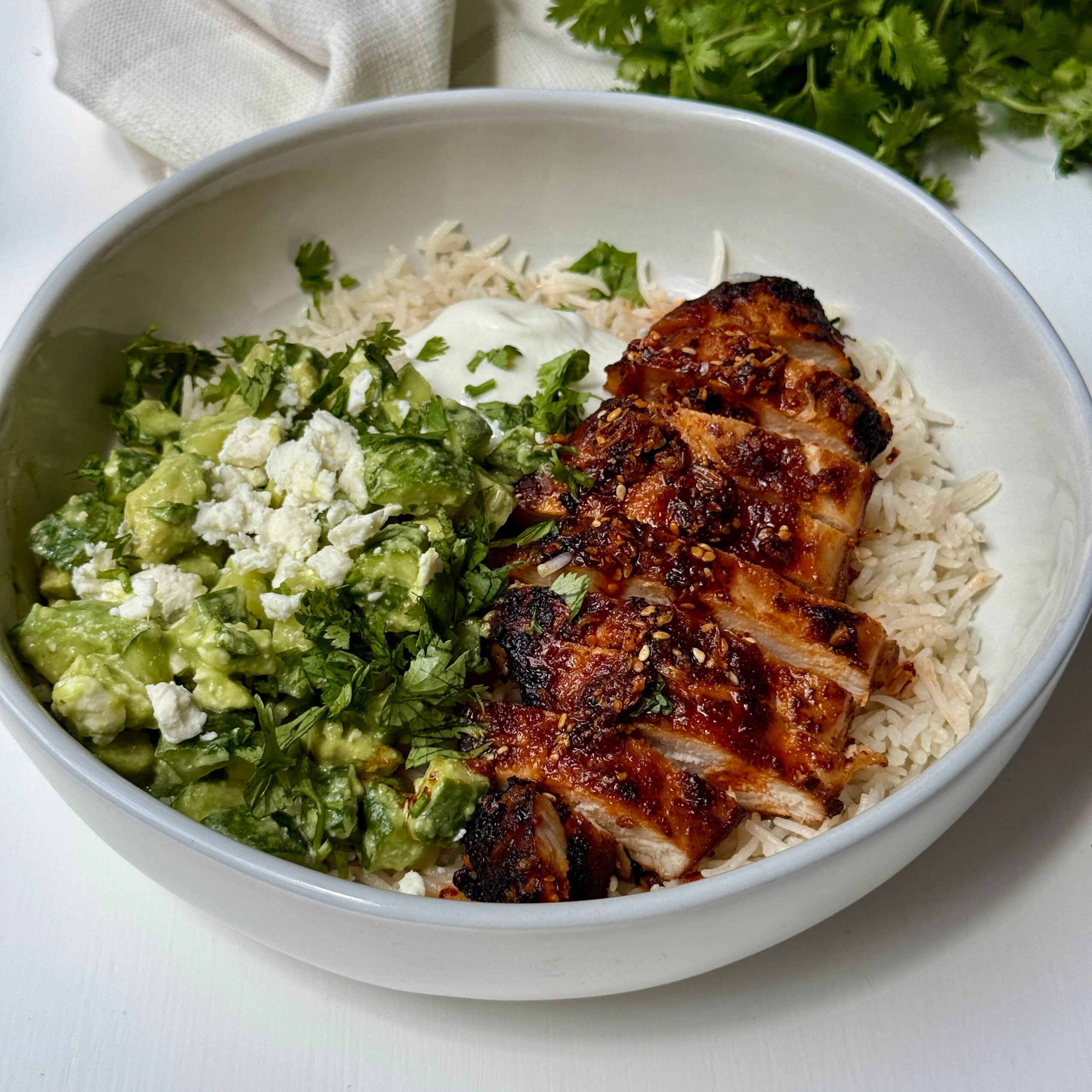 A bowl with grilled, sliced chicken over rice, served with a green avocado and feta mixture, topped with chopped cilantro and a dollop of white sauce. Fresh cilantro is visible in the background.