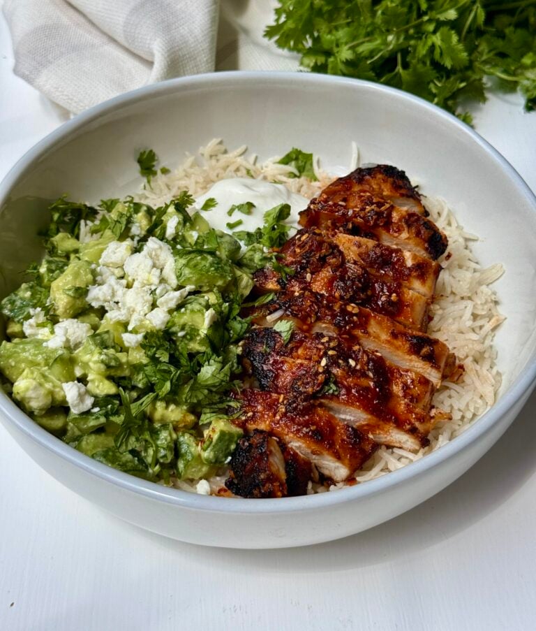 A bowl with grilled, sliced chicken over rice, served with a green avocado and feta mixture, topped with chopped cilantro and a dollop of white sauce. Fresh cilantro is visible in the background.