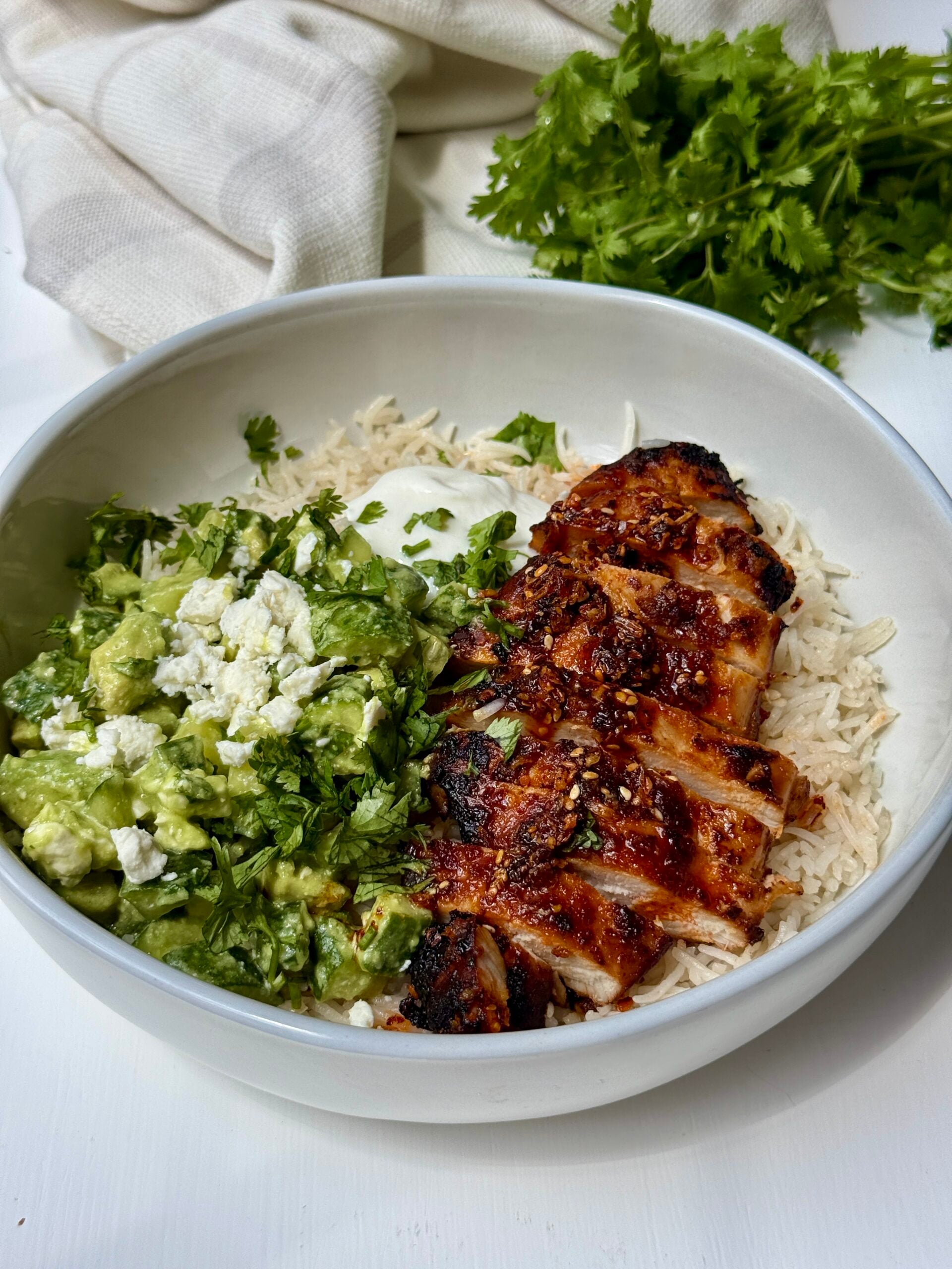 A bowl with grilled, sliced chicken over rice, served with a green avocado and feta mixture, topped with chopped cilantro and a dollop of white sauce. Fresh cilantro is visible in the background.