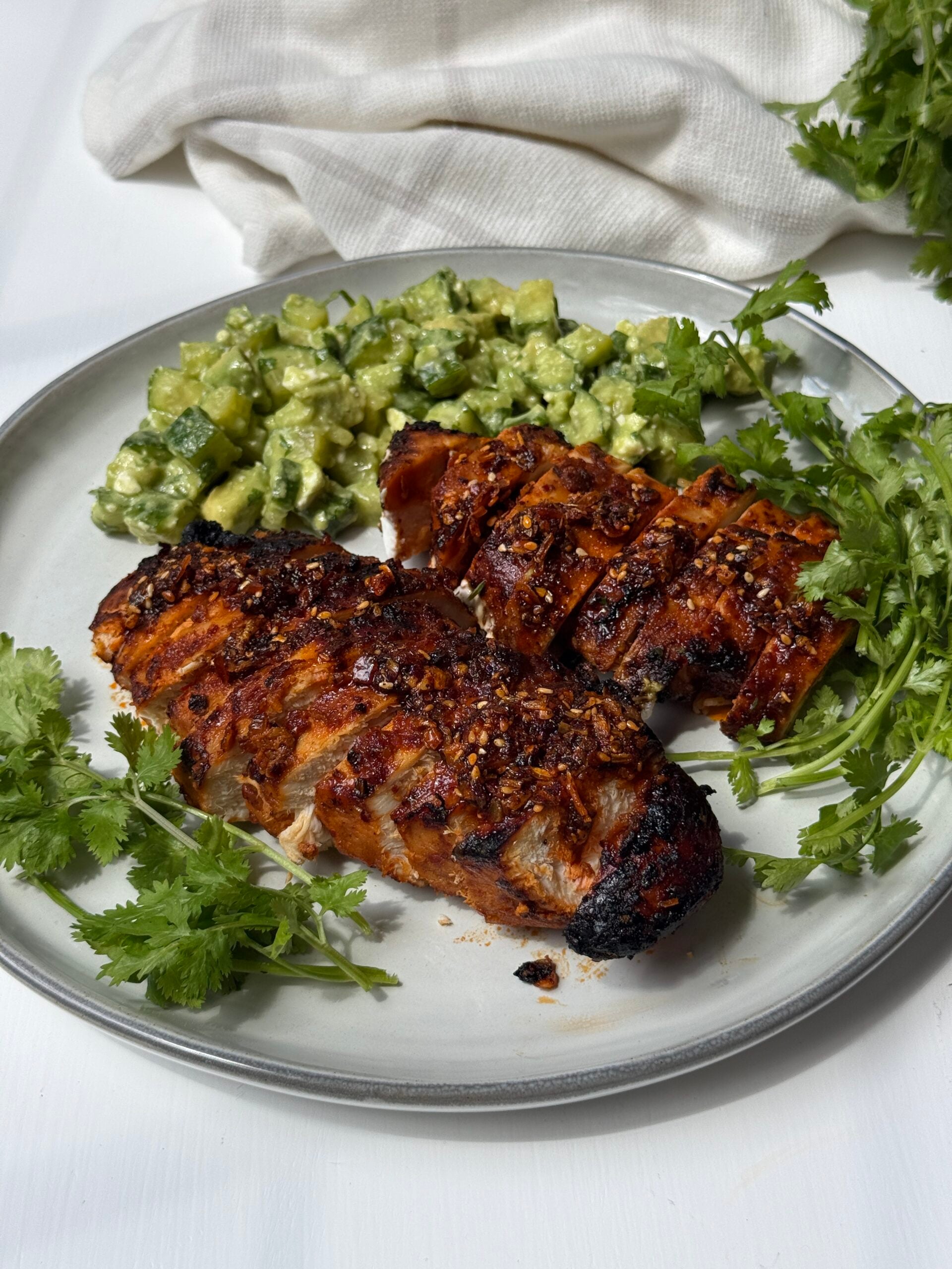 A plate with two pieces of grilled, sliced chicken garnished with cilantro, served alongside a chopped avocado salad. A white cloth napkin and more cilantro are in the background.