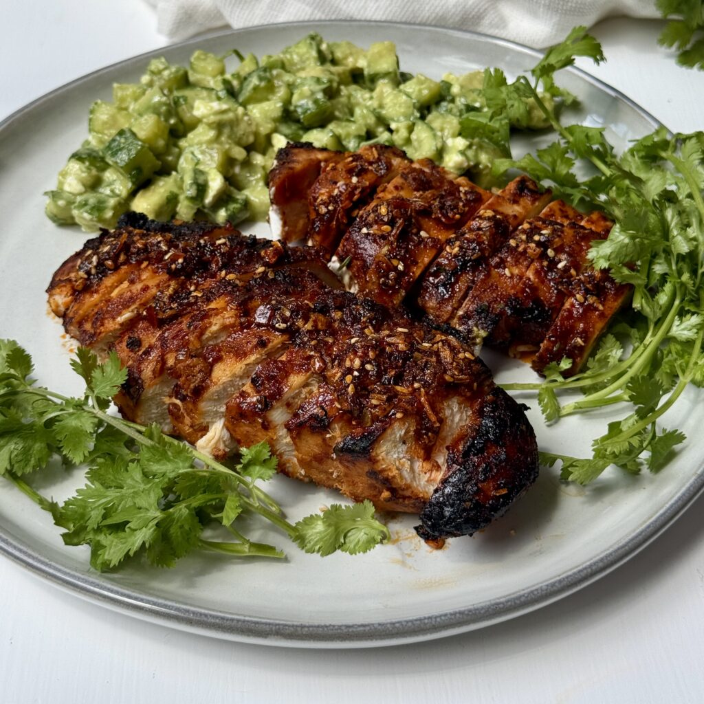 A plate with two pieces of grilled, sliced chicken garnished with cilantro, served alongside a chopped avocado salad. A white cloth napkin and more cilantro are in the background.