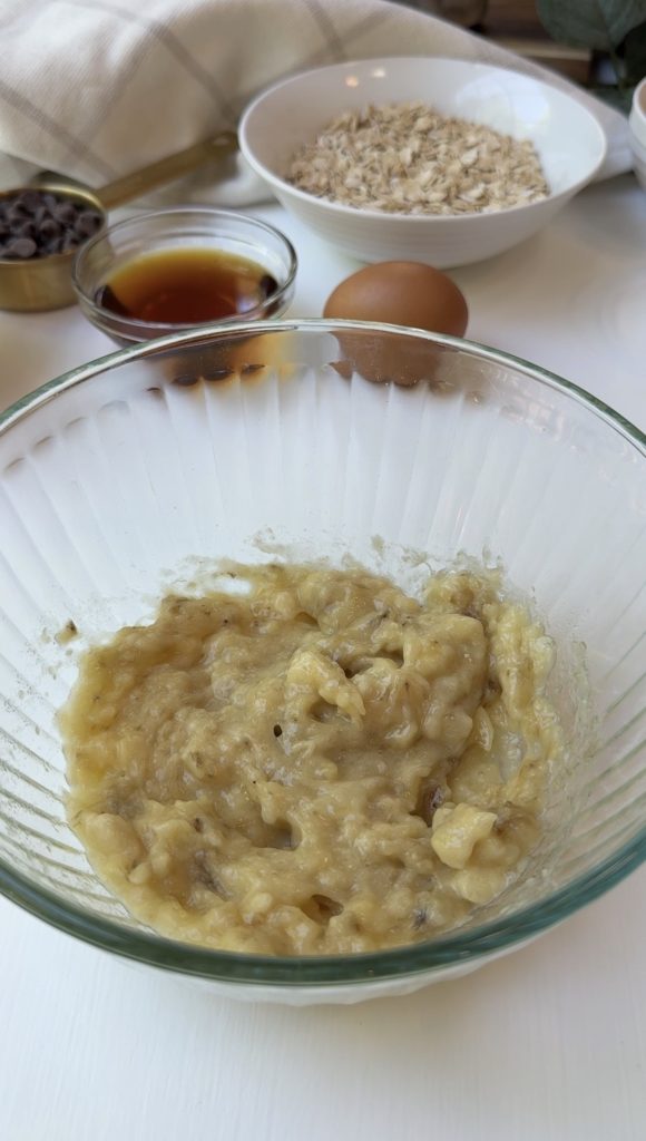 A glass bowl with mashed bananas sits on a white surface. Behind it are an egg, a glass bowl of vanilla extract, a bowl of oats, and a dish of chocolate chips.