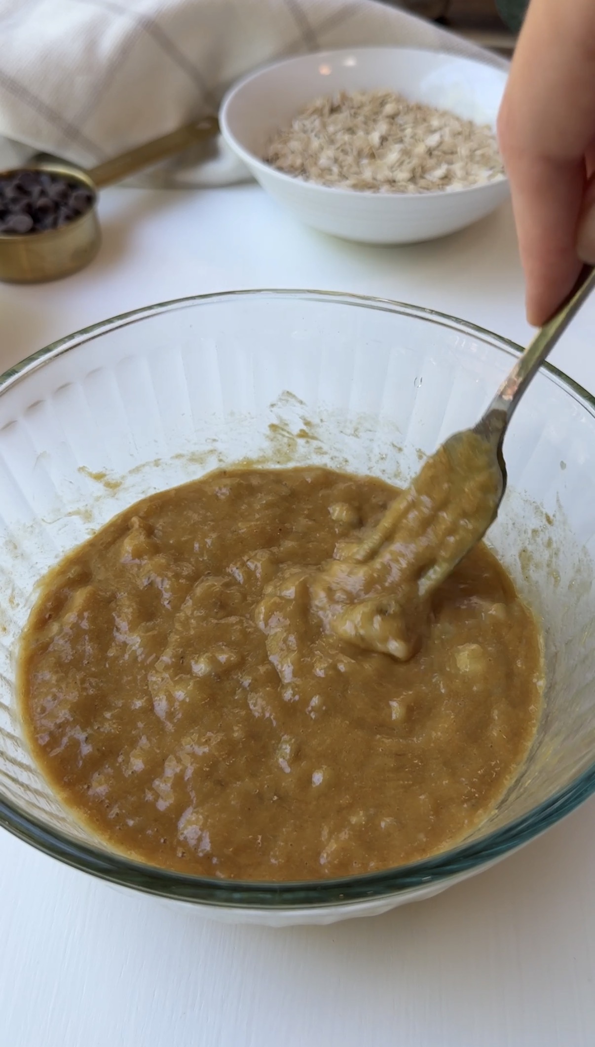 A hand uses a fork to mix a thick, brown batter in a clear glass bowl. In the background, there are measuring cups, a bowl of oats, and a white napkin on a white surface.
