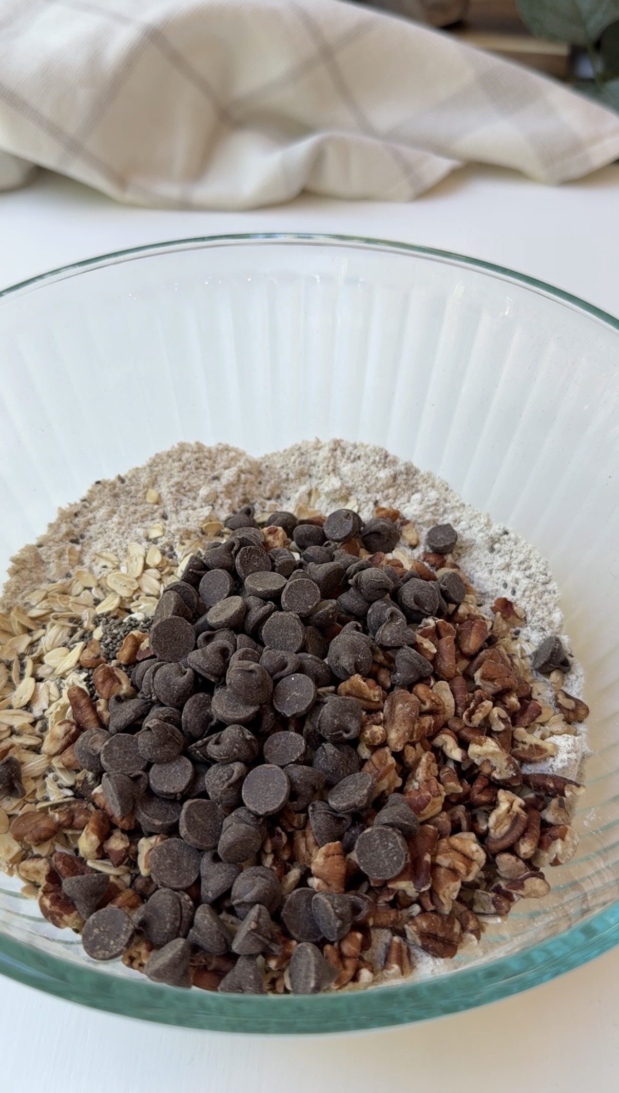 A glass bowl containing dry ingredients: flour, oats, chocolate chips, and chopped nuts, arranged in separate sections on a white surface.