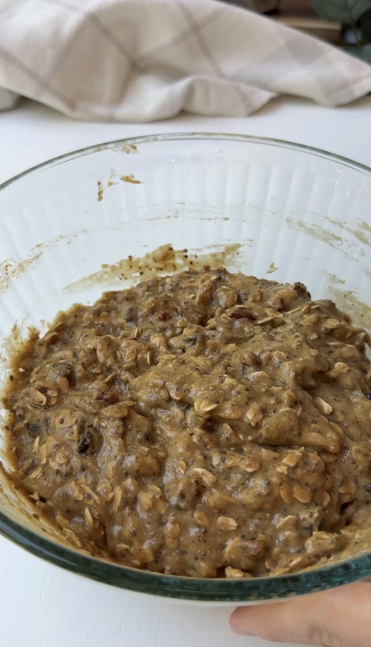 A glass bowl filled with thick, chunky oatmeal batter mixed with visible oats and seeds, sitting on a white surface. The mixture appears ready for baking or cooking.