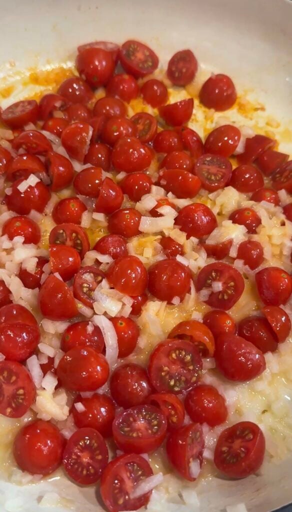 Chopped cherry tomatoes and diced onions cooking in a pan with olive oil, starting to soften and release their juices.