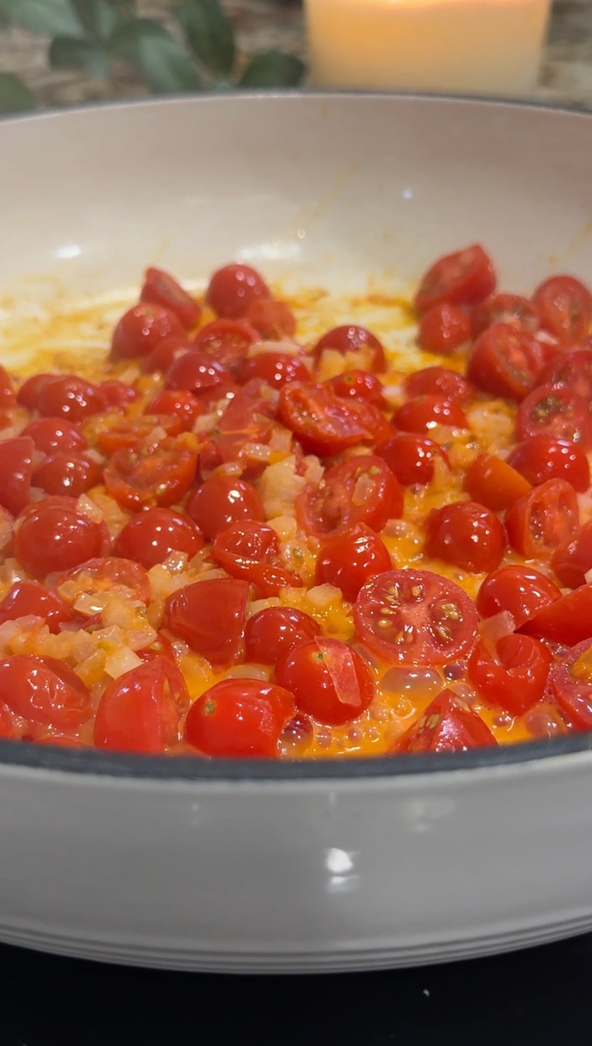 A close-up of a white pan filled with halved cherry tomatoes being sautéed with diced onions, with juices bubbling around them.