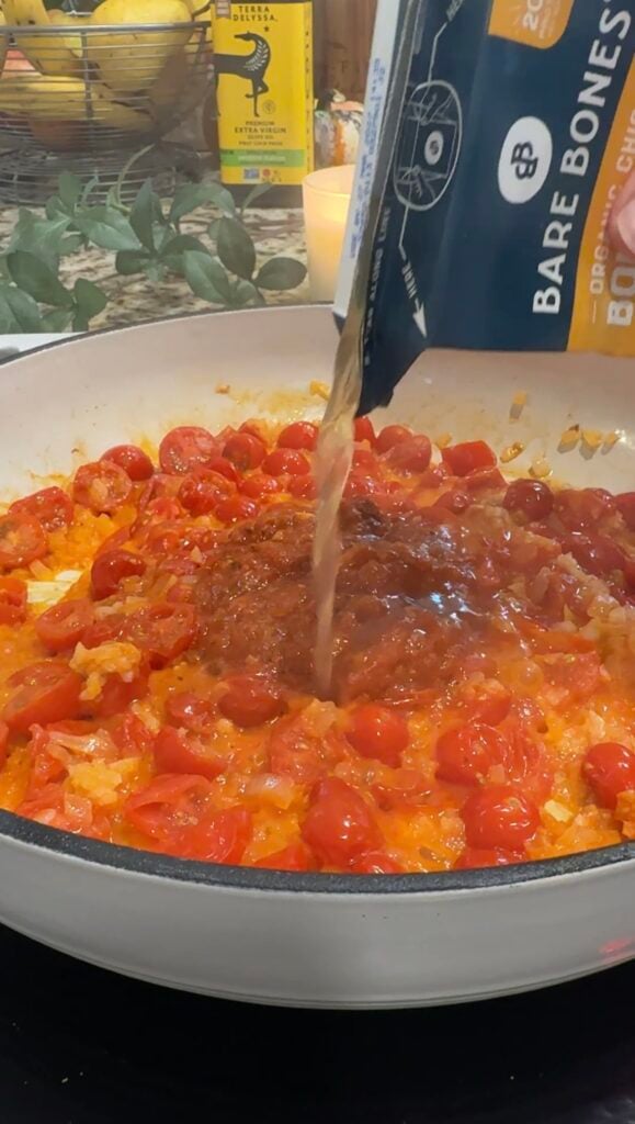 Broth is being poured from a carton into a pan with chopped onions and halved cherry tomatoes, which are being sautéed and cooked down to make a sauce.
