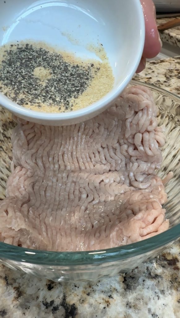 A bowl of ground turkey in a glass dish with a small bowl of black pepper and garlic powder being held above it, ready to be added.